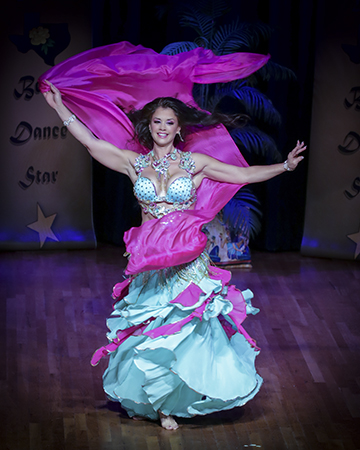 dark haired dancer wearing a pale costume smiles at the audience as she spins with a bright pink veil wrapping around her