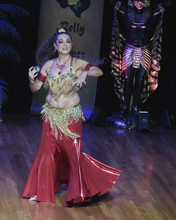 dancer wearing red costume with gold beaded bedlah smiles with playing zills during her performance