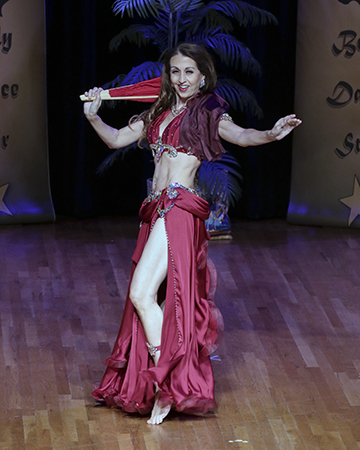 dancer wearing burgandy costume dances with a folded fan veil resting near her right sholder while her left arm is outstretched