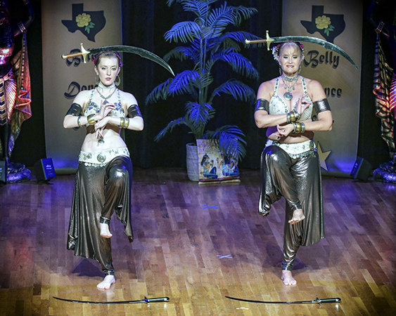 Two dancers in antique-gold liquid lame carefully balance curved swords on their heads while balancing on one foot. Additional swords are placed on the stage before them.