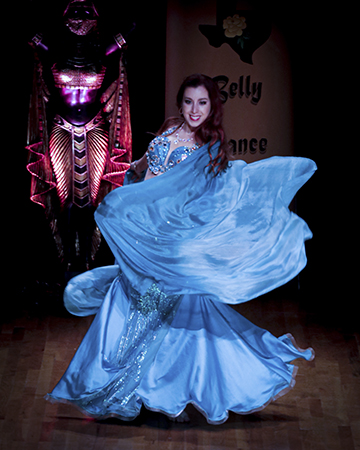 Dark haired dancer in blue swirls her veil and smiles toward the audience