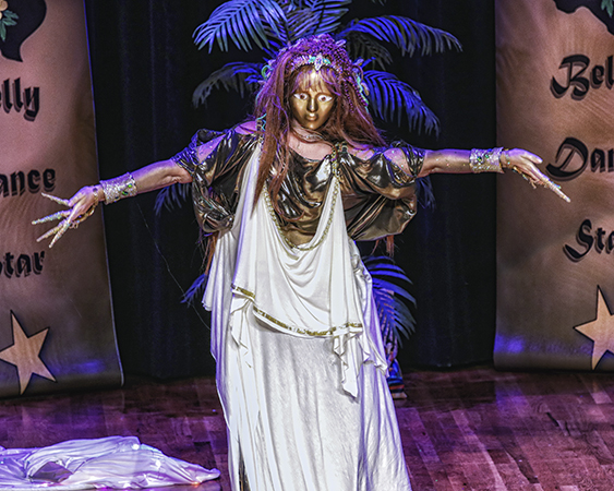 dancer in dark gold wearing a gold mask and ornate head piece, wrapped in a white layered fabric trimmed in gold