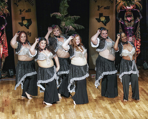 6 dancers in black costumes with sequin hip wraps and black and white striped edging, with silver bedlah and tiny black tophats, touch their hats as they pose and smile toward the audience