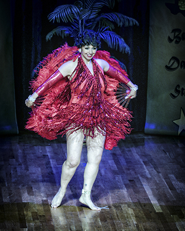 dancer wearing a fitted red costume with long, flowing fringe holds large red feather fans behind her back