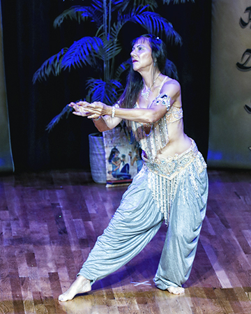 dancer wearing silver bedlah and pants holds her hands together and forward in a dramatic gesture toward her right