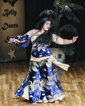 dark haired dancer in royal blue and gold dances on stage with arms outstretched