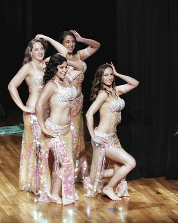 quartet of smiling dancers in blush/pink costumes and in a close group strike a pose on stage with left hands gently touching side of head