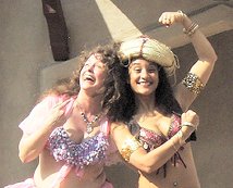 2 dancers smiling together while enjoying their time on stage, one wearing a costume hat and posing with her hand in fists and showing off her muscles