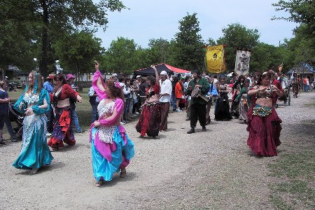 dancers, drummers, and other faire participants are shown walking down gravel path during the daily parade