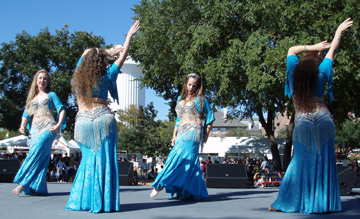 4 dancers in matching turquoise and silver costumes perform together on stage, two are facing audience and two have backs toward audience with right arms outstretched to the sky