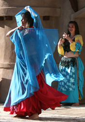 dancer wearing a red skirt wrapped in turquoise veil performs on stage with another dancer playing zills near the back of the stage