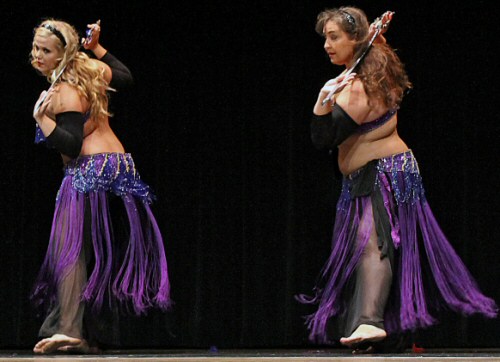 2 dancers wearing black with purple bedlah and fringe hip wraps perform with swords