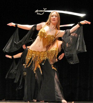 dancer wears a black costume with gold beaded bedlah and balances sword on her head during stage performance