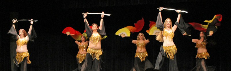 6 dancers in matching black costumes with gold beaded bedlah perform on stage, half holding silver swords overhead and the other half using fire-toned fan veils