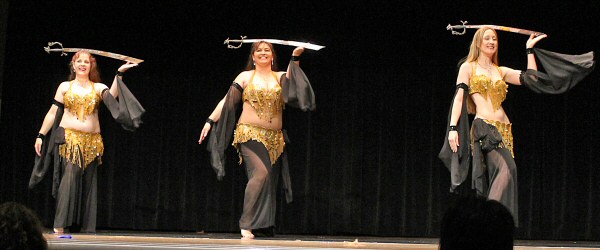 3 dancers in matching black costumes with beaded gold bedlah perform on stage with silver swords balanced on their heads