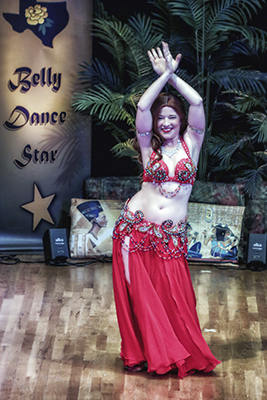 dancer wearing red costume dances smiles at audience with both arms overhead with her hands together