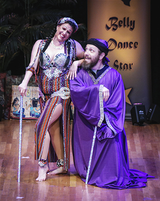 female dancer holdinga silver cane in one hand rests her arm on the shoulder of a male dancer who is kneeling on the stage and wearing a purple costume
