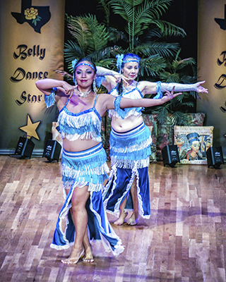 Two dancers wearing blue and silver smile at the audience as they reach their left arm to their side and right arm bends with hand just under their chins