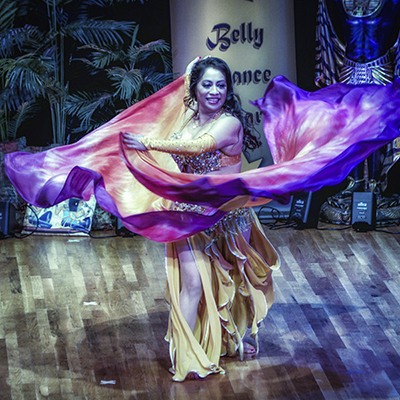 dark haired dancer wearing a warm yellow costume smiles at the audience as she spins with a pair of yellow, pink, and purple veils wrapping around her