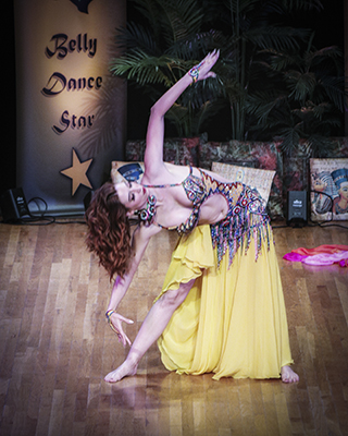 dancer in yellow skirt and multi colored bedlah bends over her right leg that is outstretched to her side