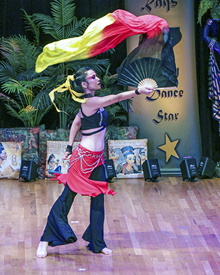 dancer wearing black pants and top with a red hipscarf and a yellow ribbon in her dark hair flutters a black, red, and yellow fan veil