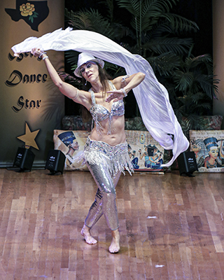 dancer wearing silver pants, bedlah, and hat flips a silver fan veil over her head while facing the audience
