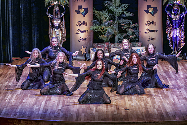 eight dancers wearing full coverage black costumes with draping sleeves kneel on both knees while facing the audience, arms outstretched at their sides