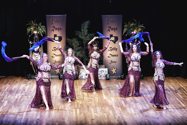 five dancers wearing matching burgandy costumes face the audience while flipping closed fan veils of purple and blue overhead